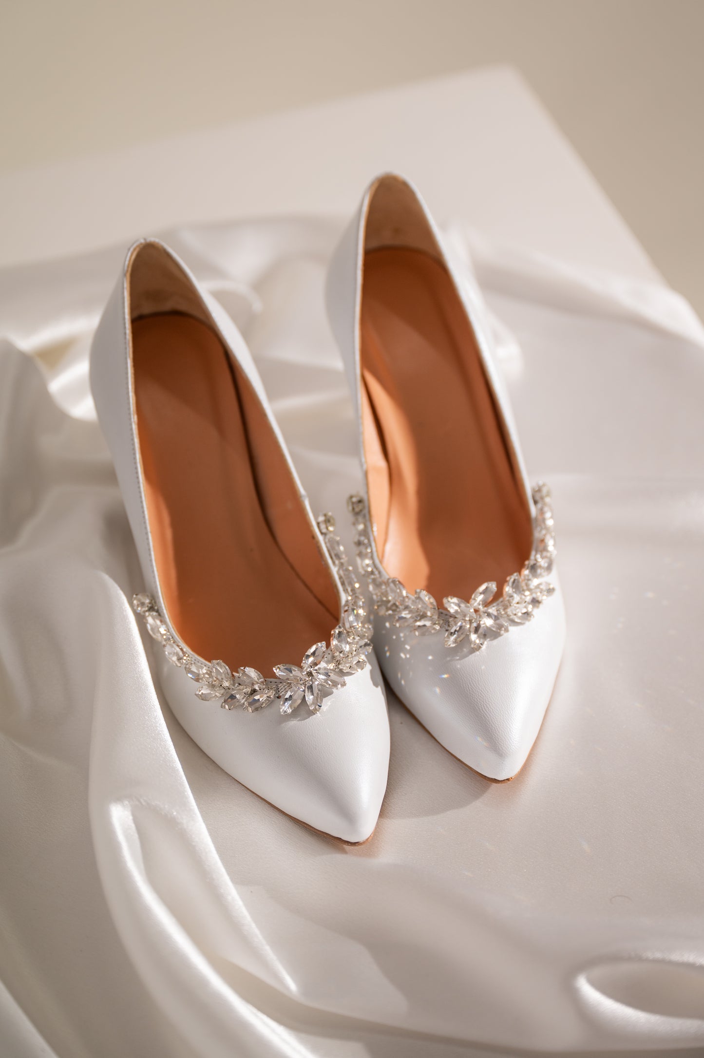 A pair of white leather bridal shoes with embellishments on the front.