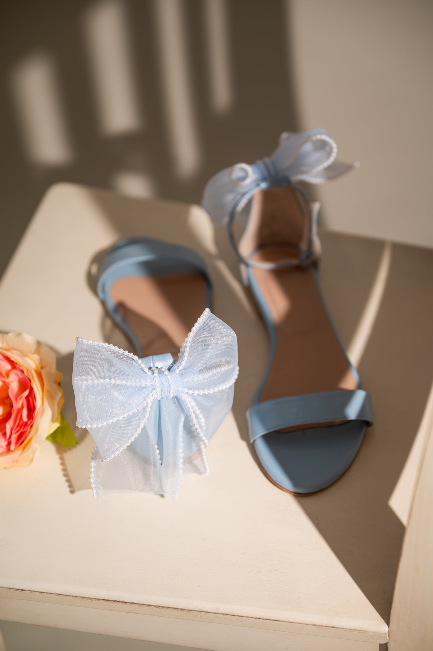 A pair of blue flat wedding sandals with ankle straps, displayed next to a decorative bow and a flower.