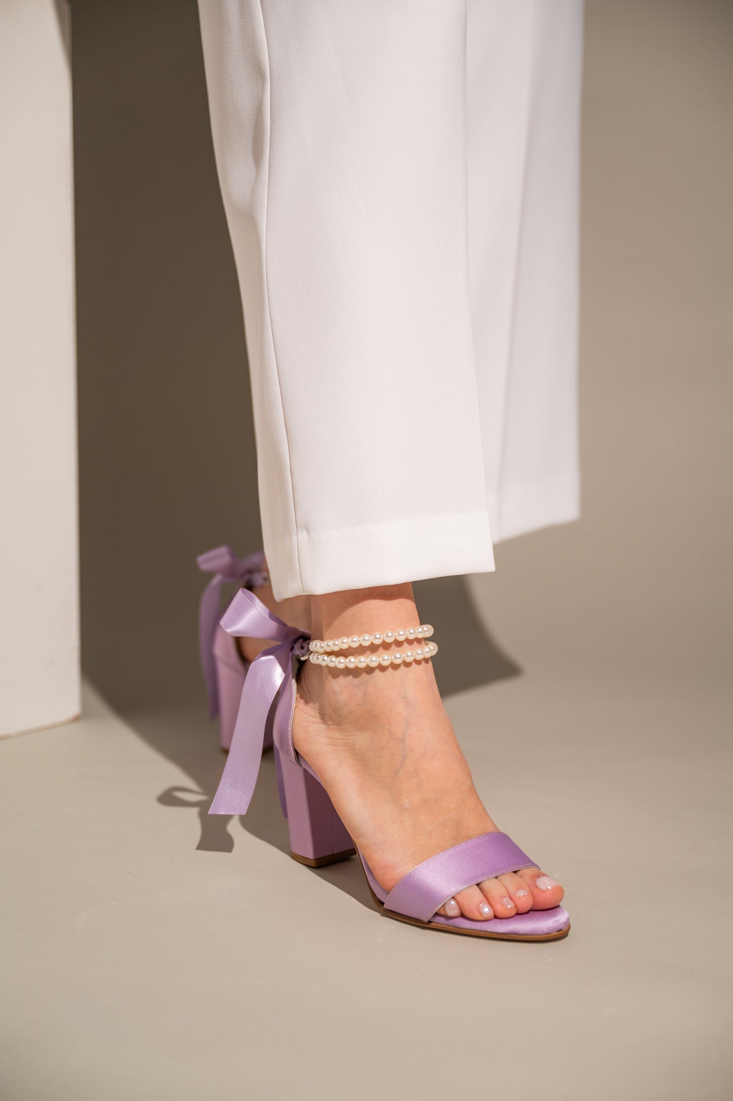 A close-up of a woman's foot wearing a lilac satin shoe with a heel, featuring a pearl ankle strap and a bow detail on the ankle.