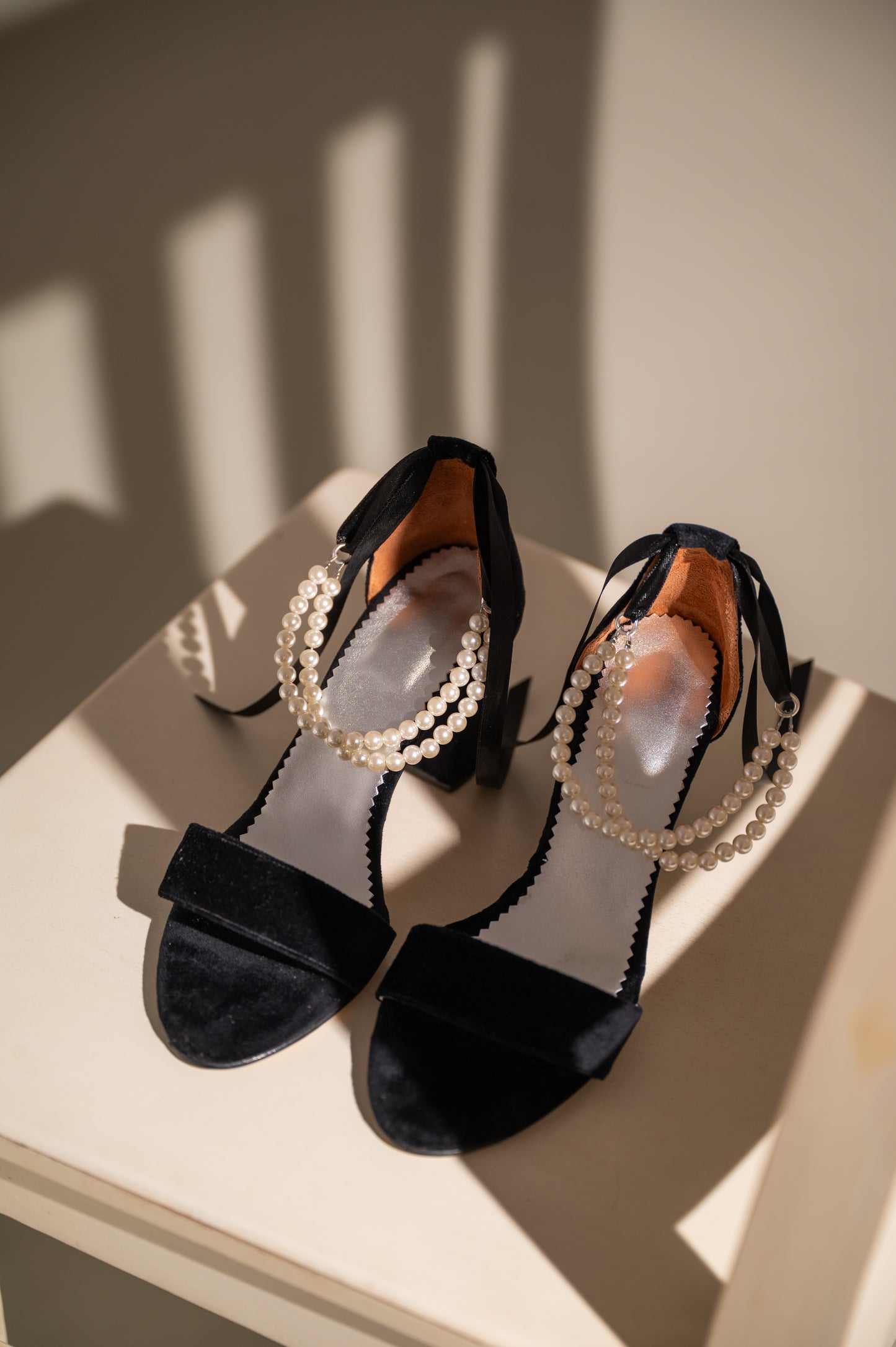 A pair of black velvet women's shoes with pearls, likely bridal shoes, displayed on a light-colored surface with a shadow falling over part of the image.