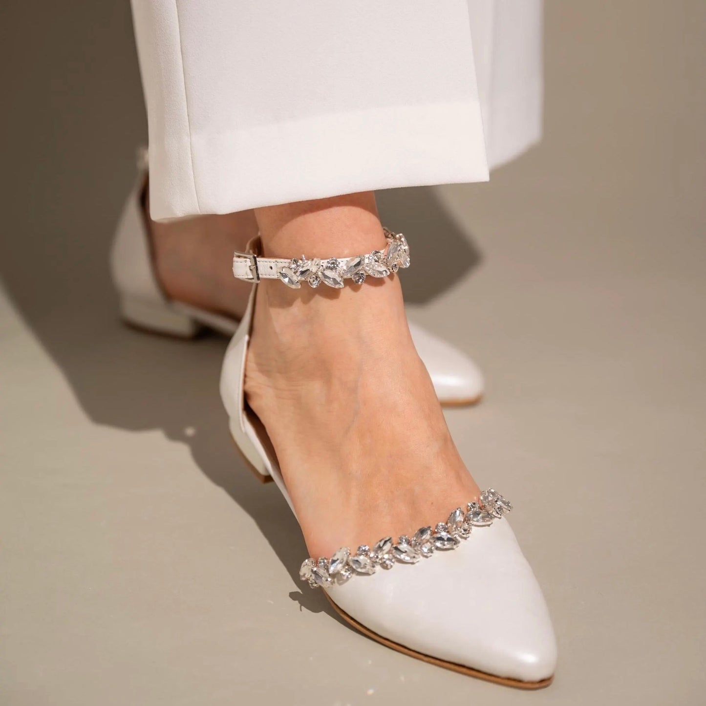 A close-up of a white flat wedding shoe with rhinestone embellishments on the ankle strap, worn by a person with their pants rolled up to reveal the shoe.
