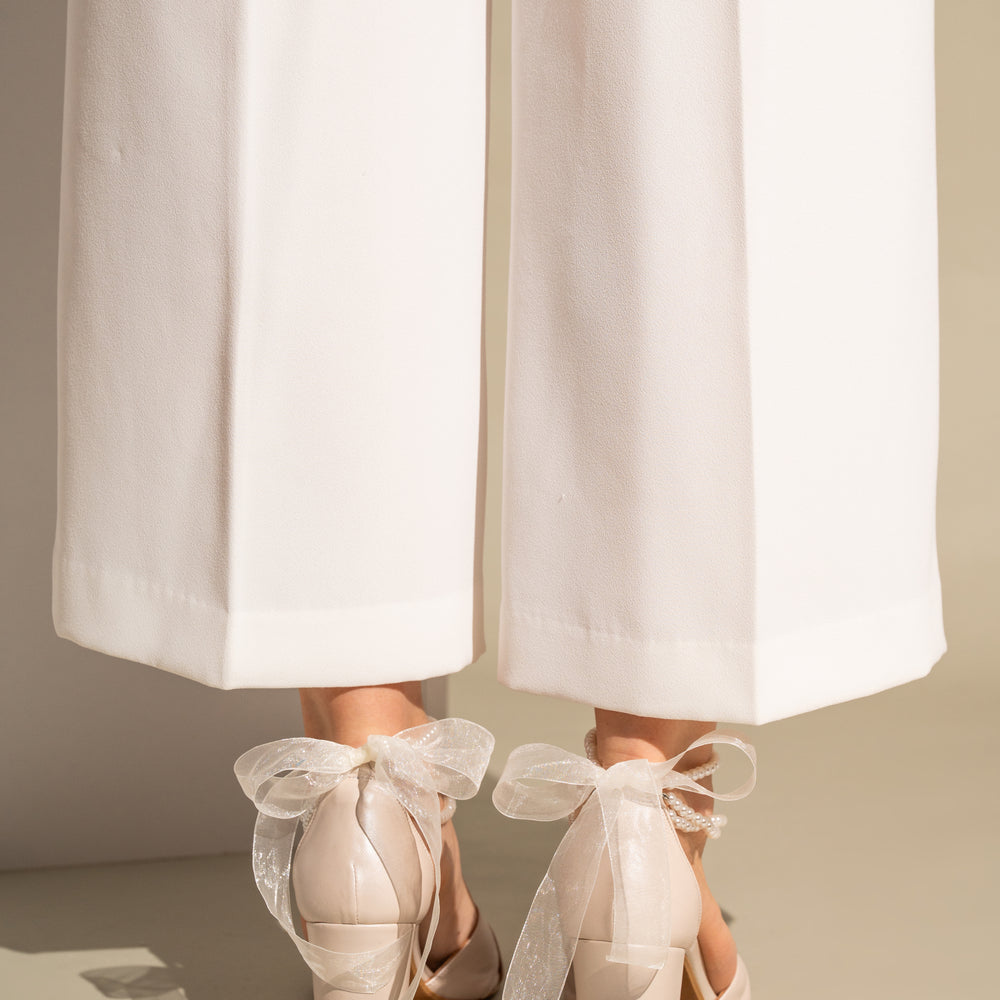 
                  
                    Ivory wedding shoes with ribbons worn with white pants on a neutral background
                  
                