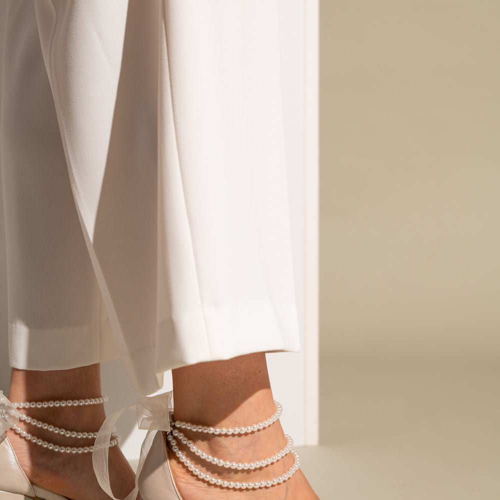 Beige high-heeled sandals with pearl straps worn with white pants on a neutral background