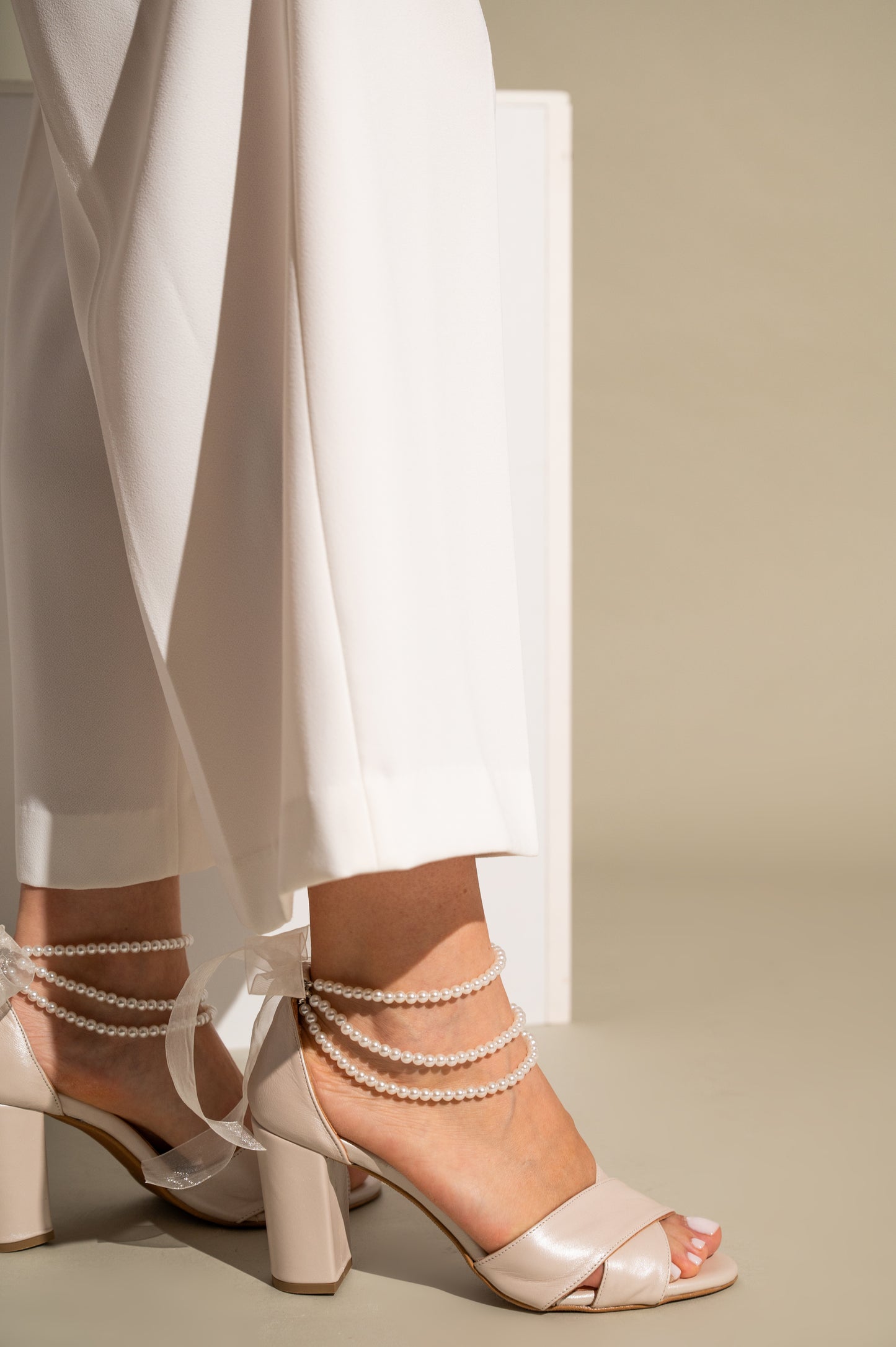 Beige high-heeled sandals with pearl straps worn with white pants on a neutral background