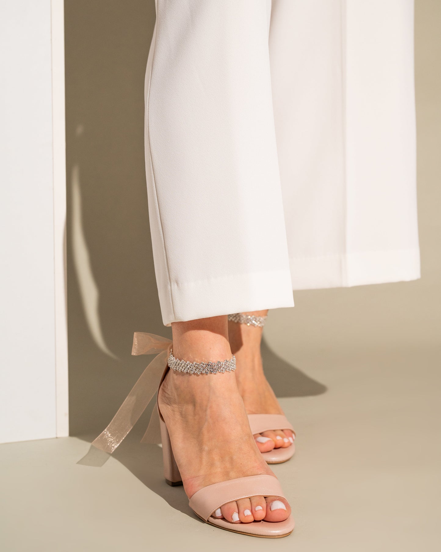 Nude high-heeled sandals with ribbon straps worn with white pants on a neutral background