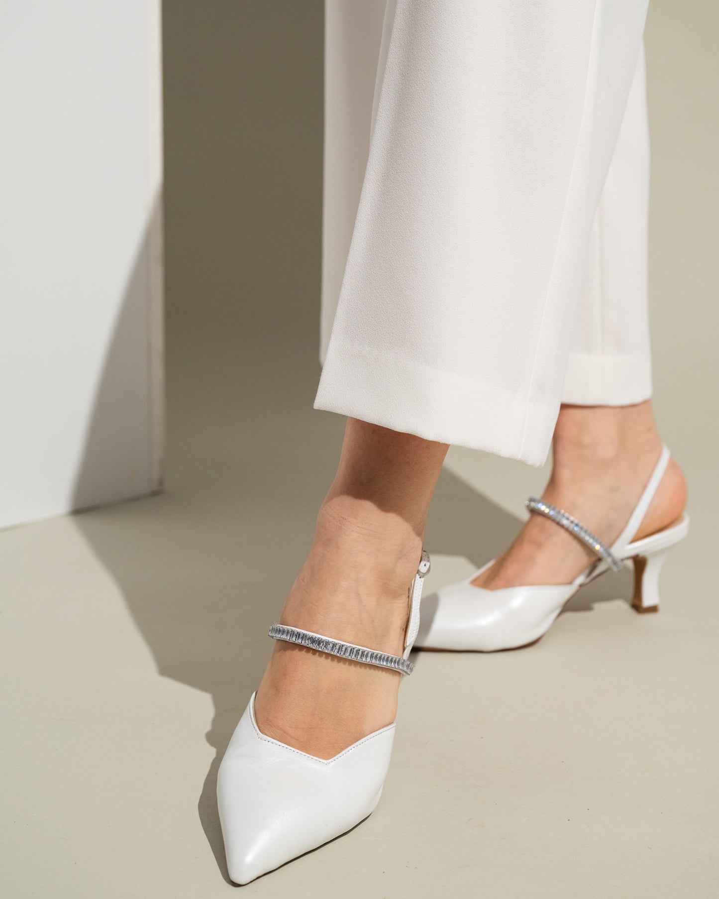 White high-heeled shoes with thin straps worn by a person in white pants on a neutral background