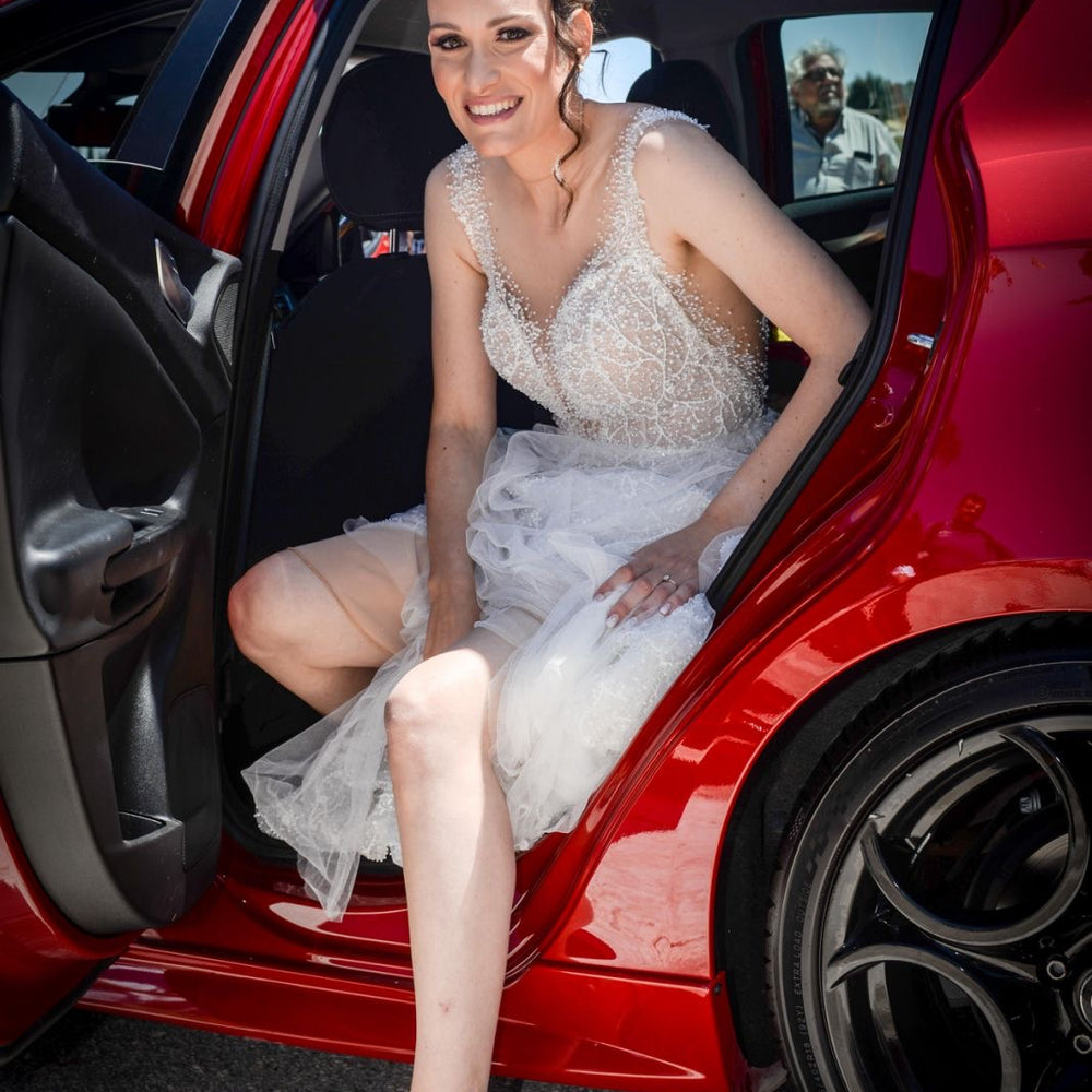 Woman in a white dress sitting in the open door of a red car on a sunny day.