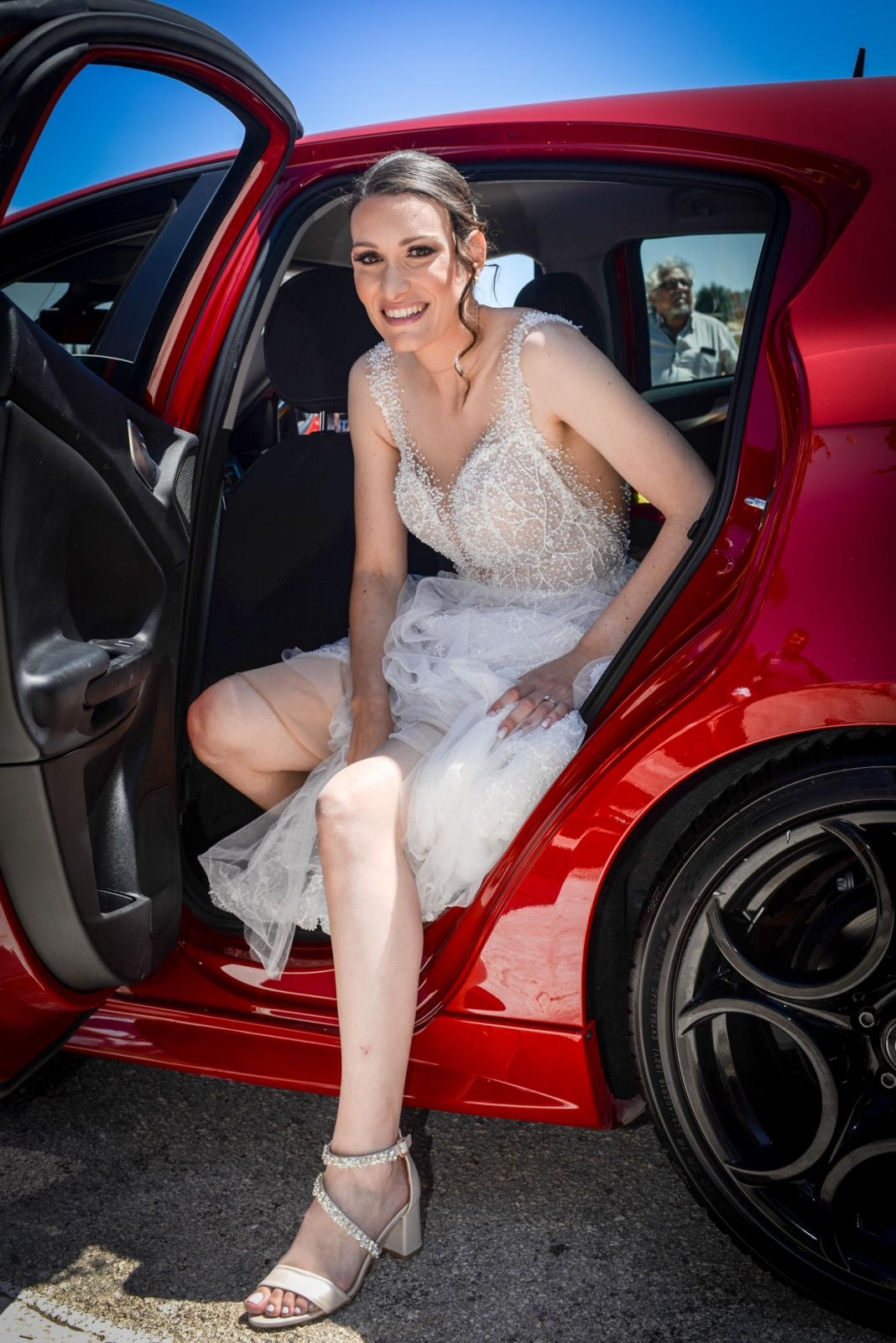 Woman in a white dress sitting in the open door of a red car on a sunny day.