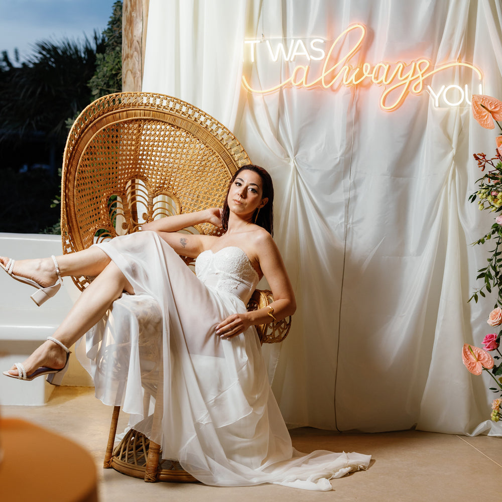 Woman in a white dress sitting on a wicker chair with a neon sign in the background.