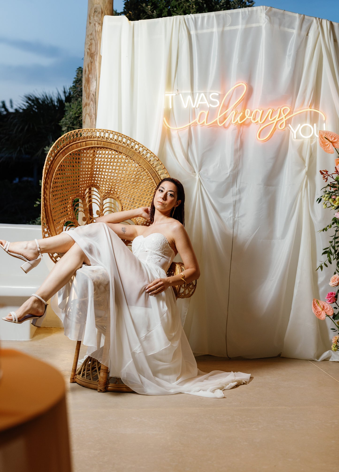 Woman in a white dress sitting on a wicker chair with a neon sign in the background.