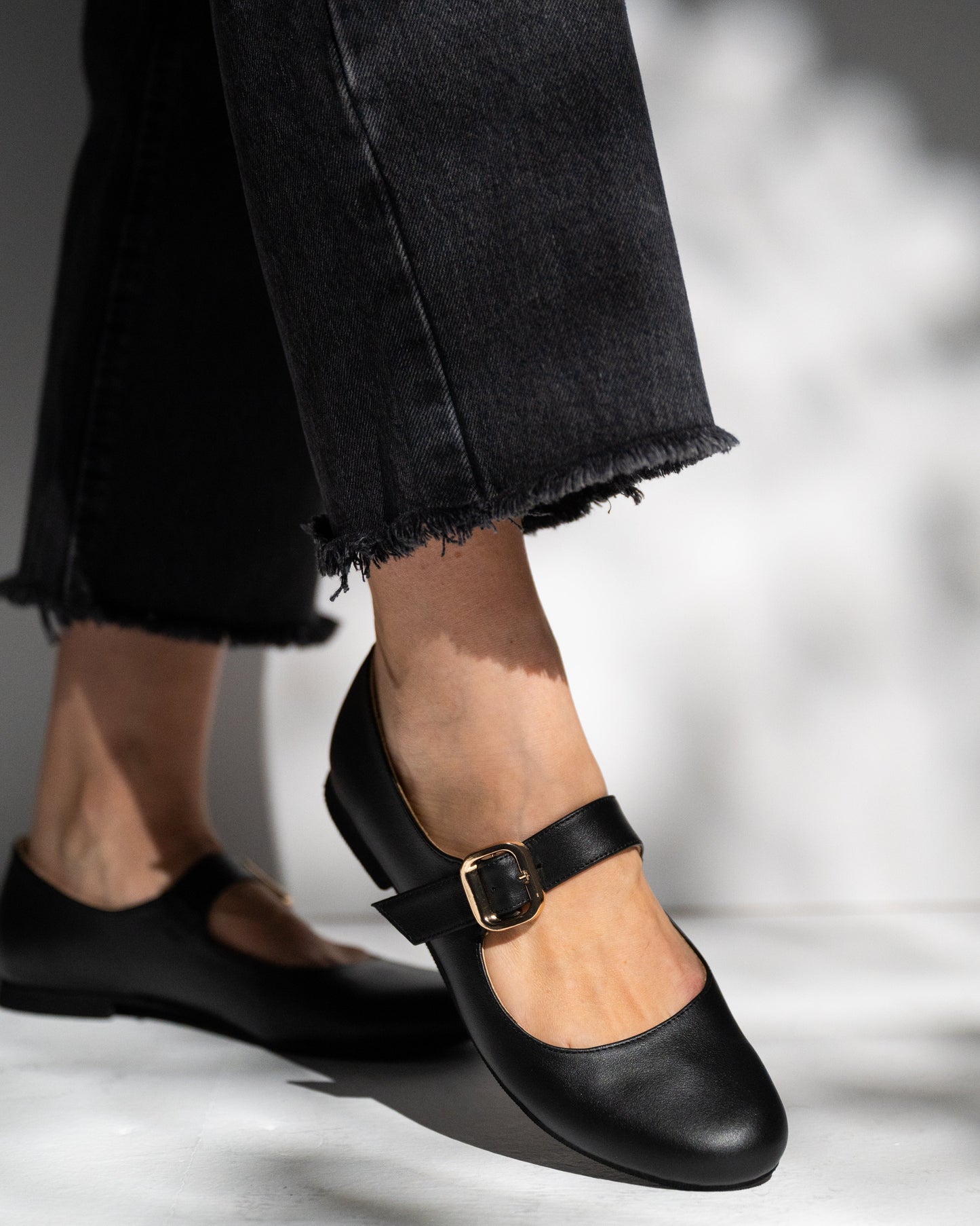 Black flat shoes with a buckle worn with black jeans on a light background