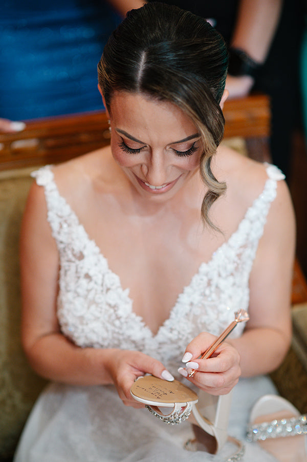 Woman in a white dress holding a compact mirror and makeup brush.