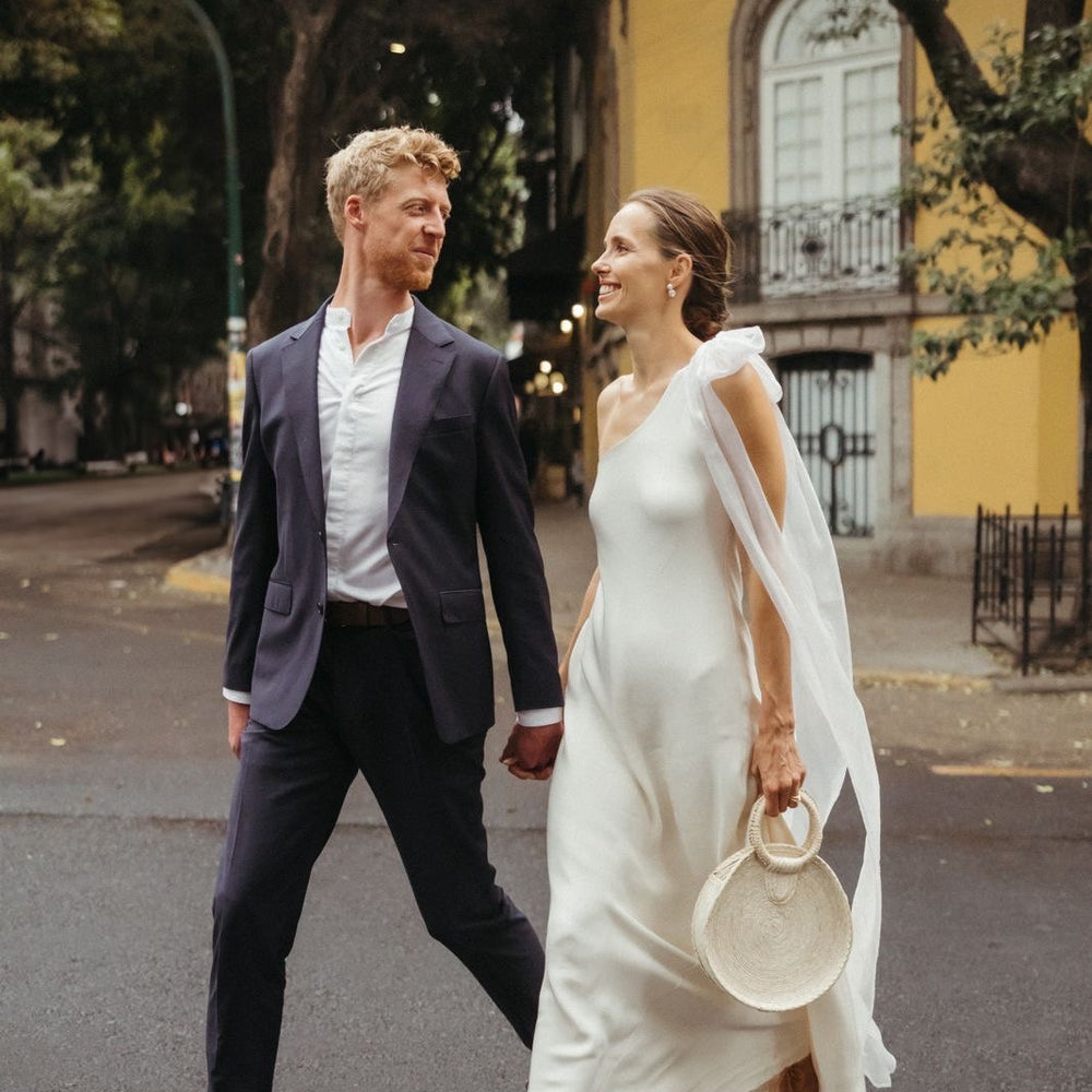 Man and woman in wedding attire walking on a street with a yellow building in the background