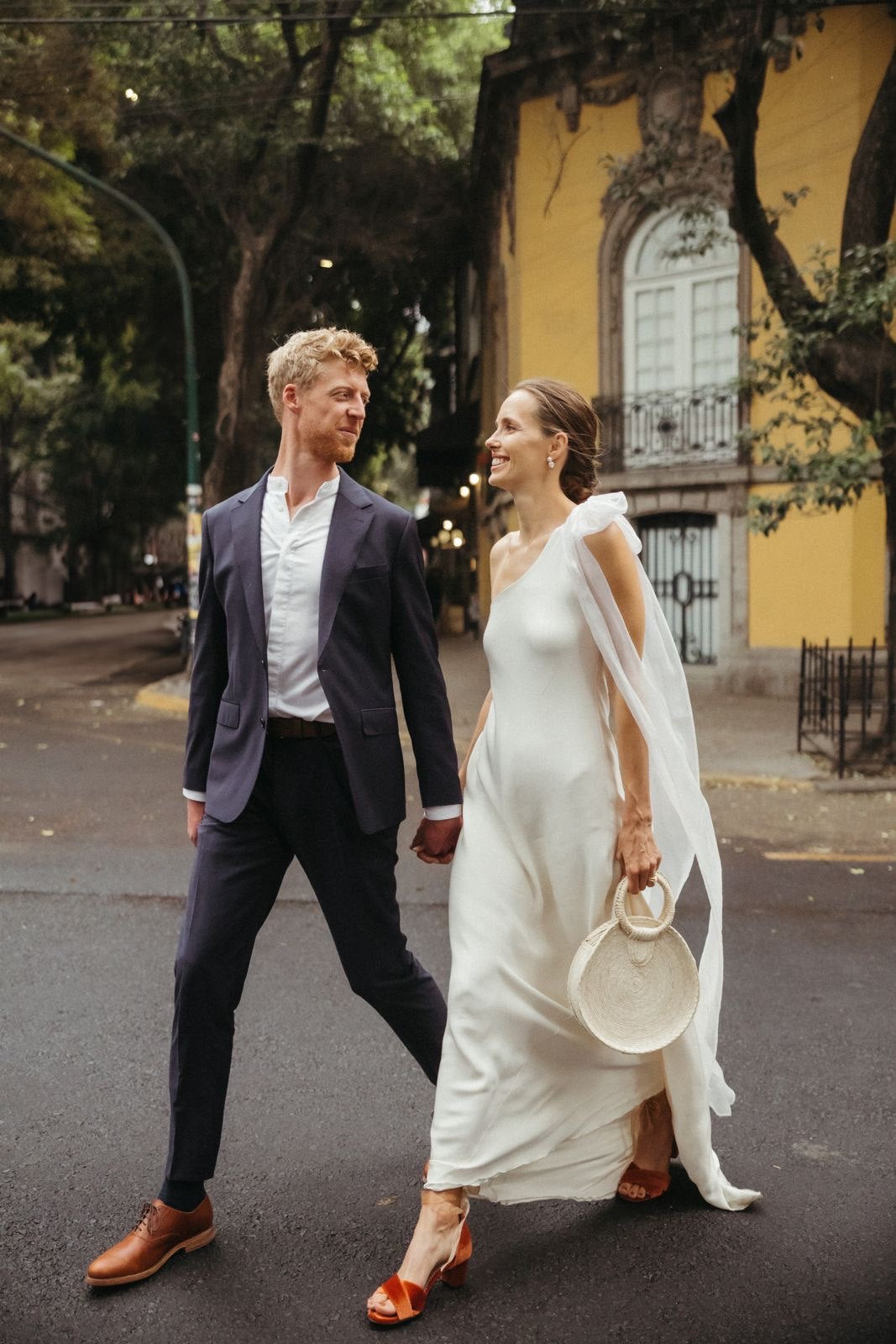 Man and woman in wedding attire walking on a street with a yellow building in the background