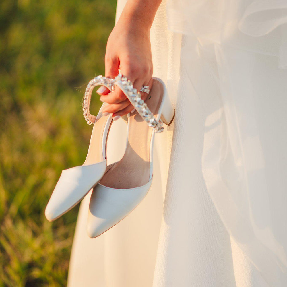 White high-heeled shoes with decorative straps held by a person in a white dress against a blurred natural background.