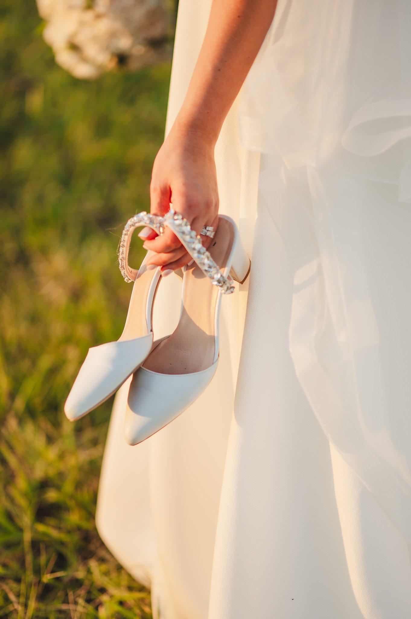 White high-heeled shoes with decorative straps held by a person in a white dress against a blurred natural background.