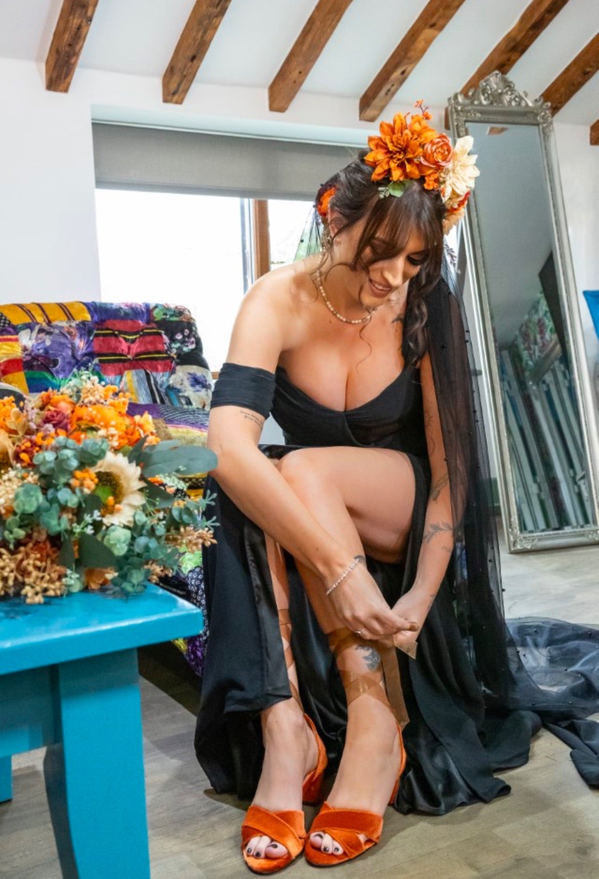 Woman in black dress and orange sandals sitting on a blue stool with flowers and a mirror in the background.