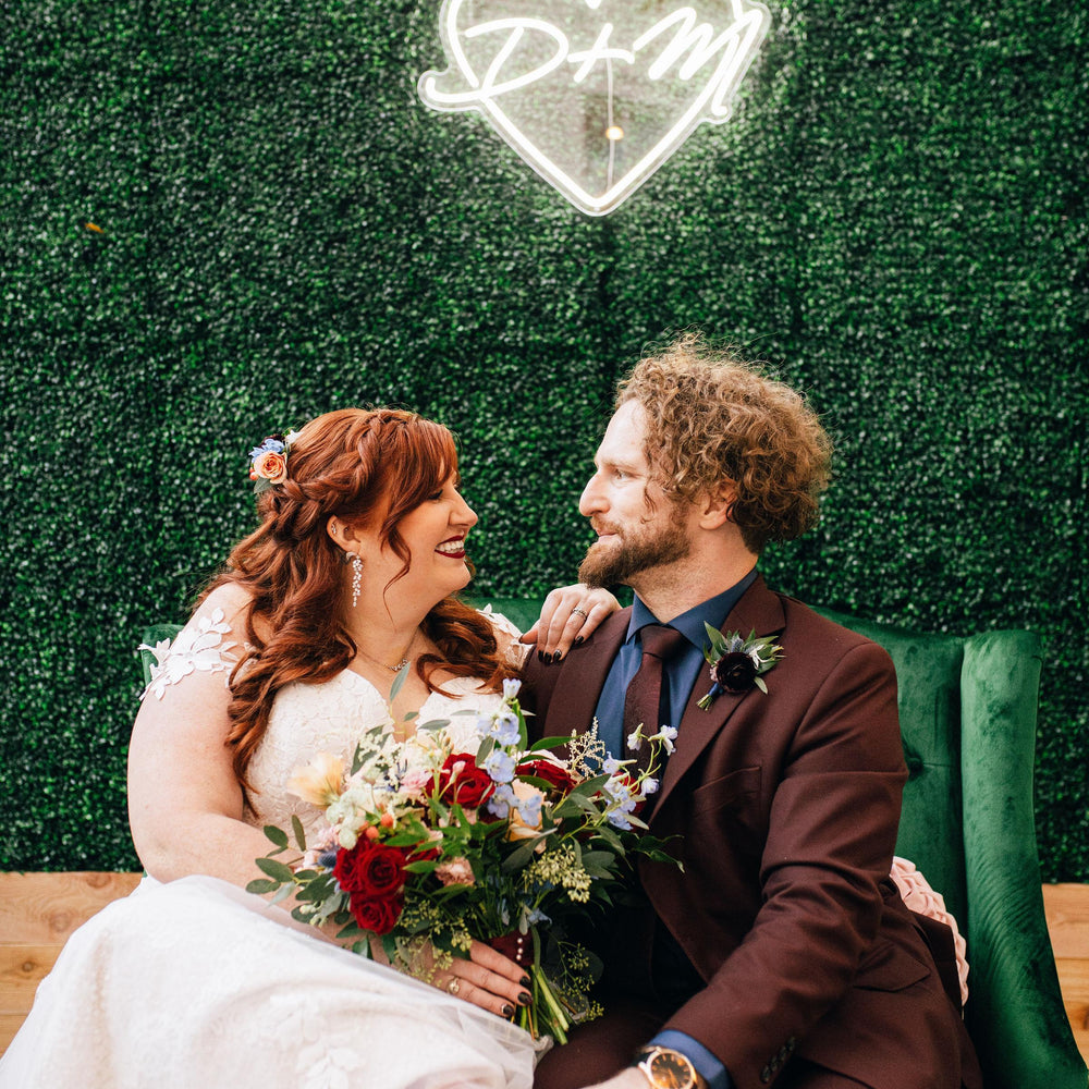 Couple in wedding attire sitting together with a decorative heart sign in the background.