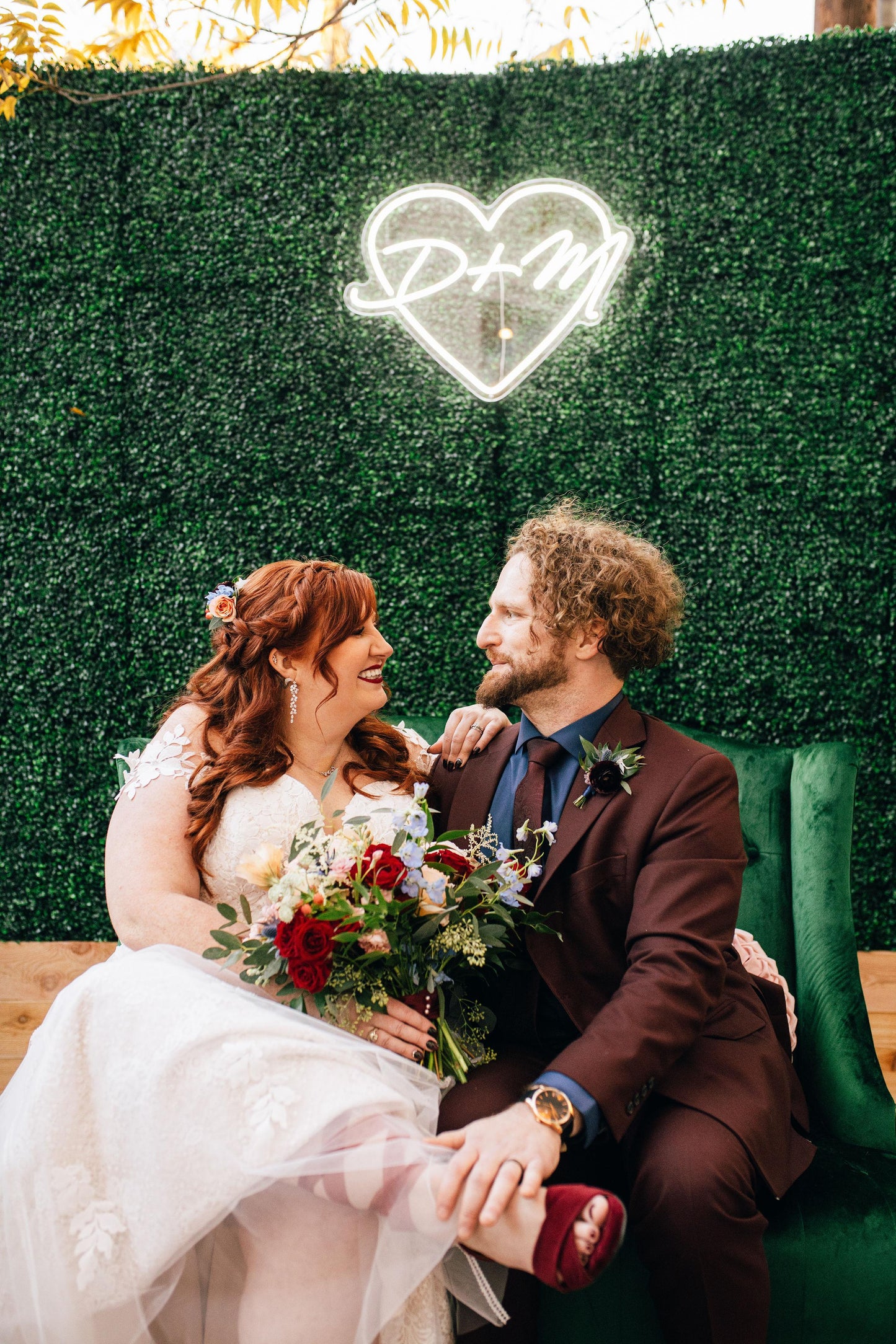 Couple in wedding attire sitting together with a decorative heart sign in the background.
